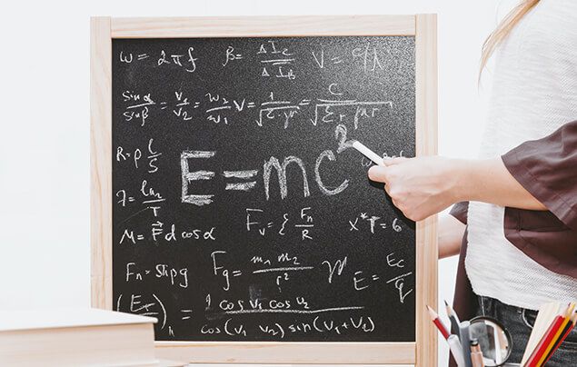a chalkboard with lots of different mathematical equations on it. one larger equation is in the centre, it reads E = m c-squared. A white hand holding a piece of chalk is pointing to this equation