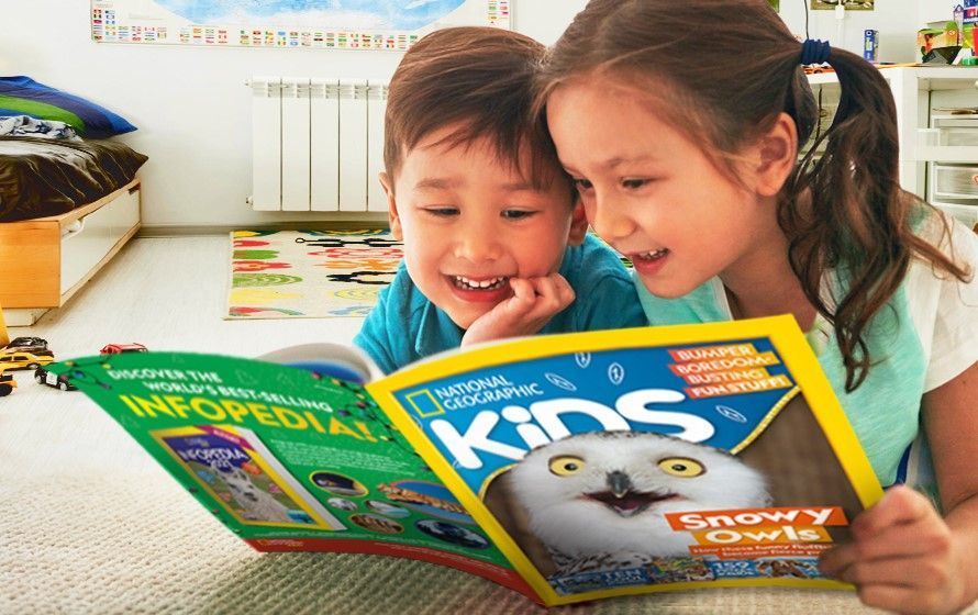 two kids are lying on the floor, holding a copy of national geographic kids magazine between them. They're reading together and smiling.