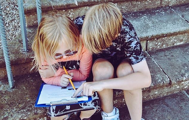 two blond children, a boy and a girl, sit together. they are filling in a clipboard and clearly working together.