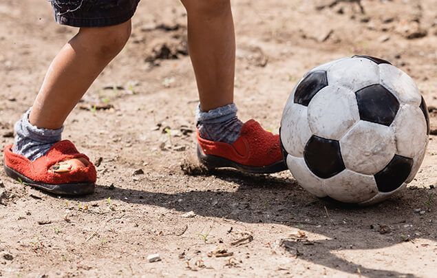 a child kicks a football on dusty ground. they are wearing slippers on their feet rather than trainers. 