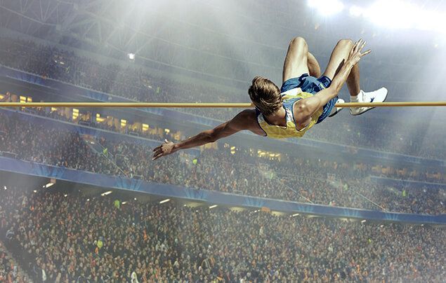 a man just crosses the bar of a high jump. behind him, a crowd of thousands cheers from a stadium