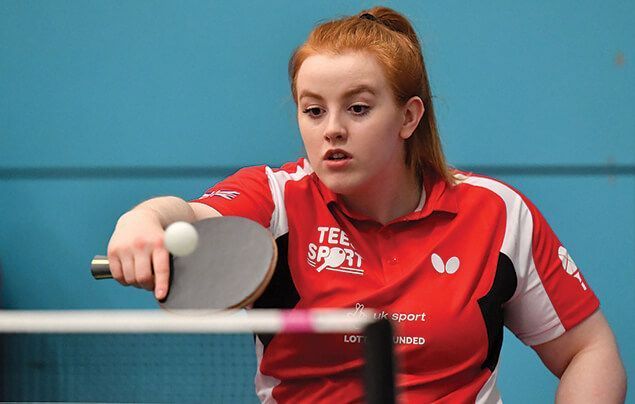 Megan looks focused as she stretches to hit a small table tennis ball with her paddle