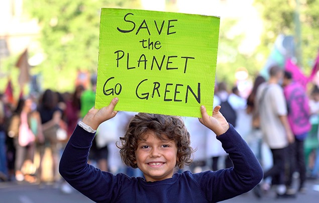 eco-anxiety | a young boy is smiling at the camera and holding up a green sign that reads 'save the planet! go green'. Behind him, a crowd of people have gathered with their own signs; they are at a protest. 