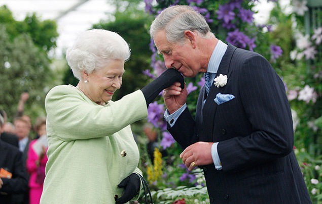 King Charles III | Charles, wearing a dark suit and a white rose in his lapel, kisses the hand of his mother Queen Elizabeth, who is wearing a pal green jacket and black gloves. They are surrounded by greenery, at a flower show.