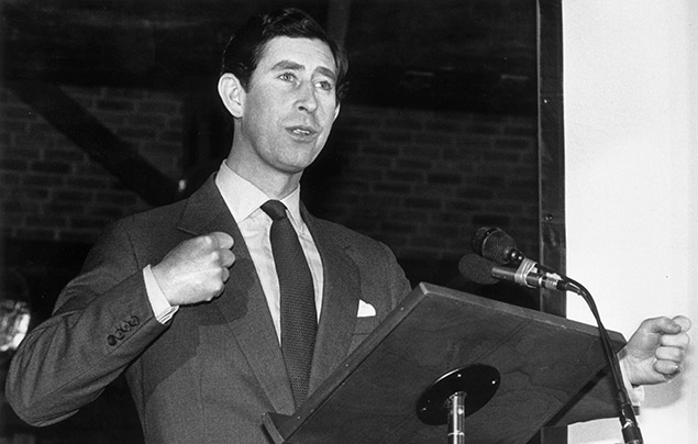 King Charles III | In this black and white photograph, a young Charles stands behind two microphones and gives a speech. He is wearing a black suit and looking earnestly into the crowd.