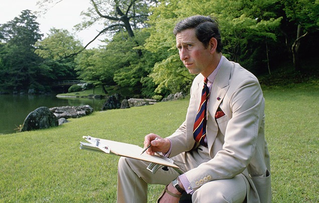 Charles sits with a sketchpad on his knee. He's outside, with trees behind him, and wearing a cream suit. He is sketching with a pencil.