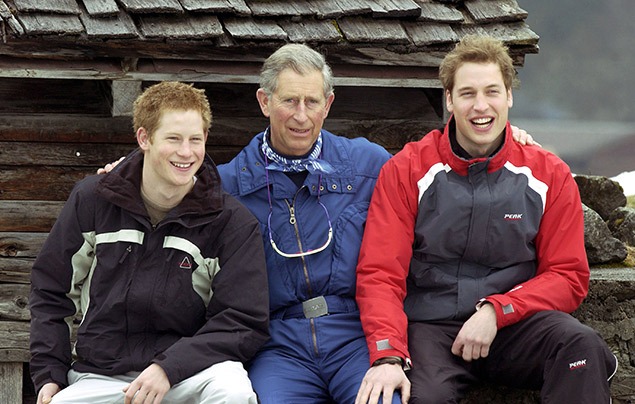 King Charles III | King Charles sits outside a ski hut. Prince William is sat to his right, and Prince Harry to his left. They are all smiling at the camera.