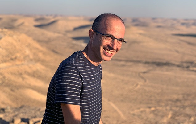 yuval noah harari stands in frond of a sandy desert. He wears a striped t-shirt and glasses and is smiling at the camera.