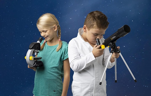 wo kids against a starry background, one dressed in green and holding the microscope, one dressed in white and holding the telescope, which rests on a small tripod. 