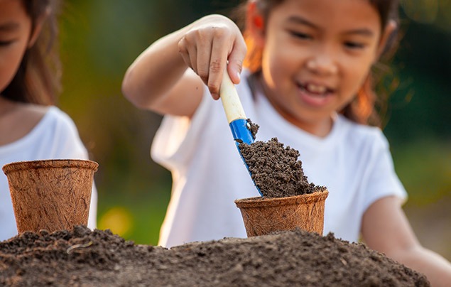 how to compost | a small girl in a white t-shirt uses a little trowel to put soil into a fibre plant pot, ready to plant in the ground