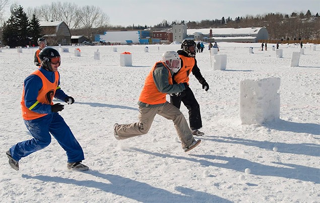 three teenagers wearing bright orange bibs and thick snow gear run across a makeshift 'pitch' on the snowy ground. They are all wearing helmets, and their hands are full of snowballs.