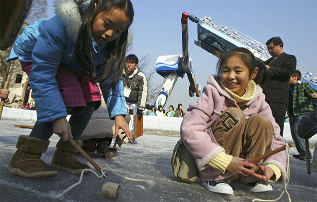 two young girls crouch down on an icy floor. They each hold a stick with a string attached to it in their hand, and a small wooden top sits in front of them on the ice.