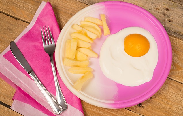on a pink plates sits a blob of white yoghurt with half an orange apricot sat in it like an egg yolk. beside it are some slices of apple cut up to look like chips.