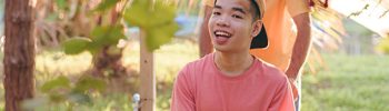 communicate better | a father pushes his son in a wheelchair towards the camera. They are outside on a grassy path and walking beneath a tree. The son is smiling broadly. They are both wearing caps; the son's is backwards on his head.