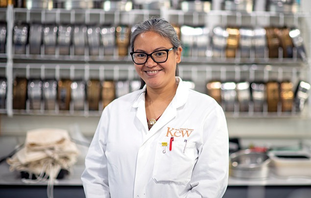 Dr Aisyah Faruk has long grey hair and wears large black glasses. She has a white lab coat on and stands in front of a row of metal pans used to clean seeds