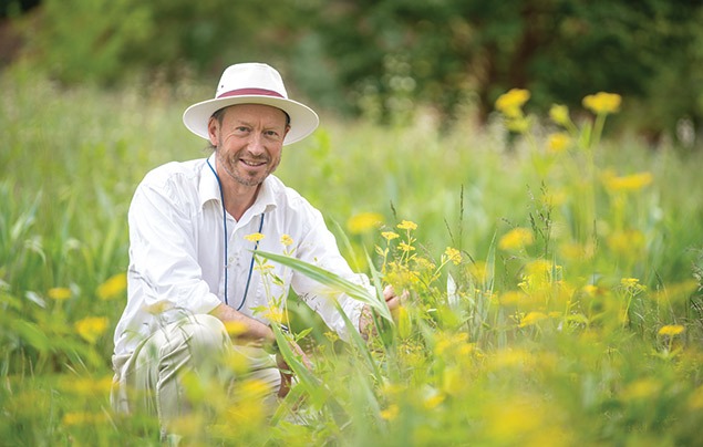 Michael Way is a white man with a stubbly beard. He's wearing a white shirt and wide-brimmed hat as he crouches in a green field, holding a yellow-flowered plant that's growing from the ground and smiling at the camera