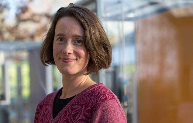 anne visscher is a white woman wearing a red jumper and standing in a bright corridor. She has chin-length brown hair and brown eyes and is smiling into the camera.