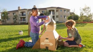 Two children outdoors assemble a cardboard rocket ship using tape and foil. Sitting on grass in a sunny park, they channel The Eurekas spirit of discovery. Apartment buildings and a helmet rest in the background.