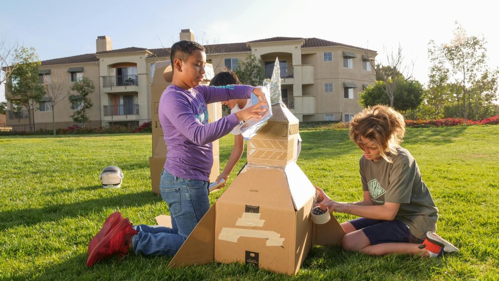 Two children from The Eurekas build a cardboard rocket ship on a grassy lawn, using tape and aluminum foil. An apartment building and trees are in the background, while sunlight shines over their imaginative scene.