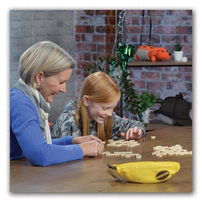 Two people, an adult and a child, sit at a table smiling and playing Bananagrams. A yellow game pouch is on the table, with plush toys and decorations in the background.
