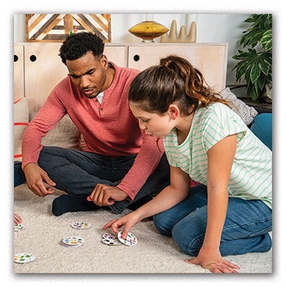 A man and a young girl sit on a carpeted floor, focused on playing Dobble. Several circular cards are spread out between them in a casual home setting with plants and cabinets in the background.