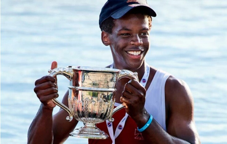 A smiling Radley rower in athletic gear and a cap holds a large silver trophy with both hands, standing proudly in front of a body of water.