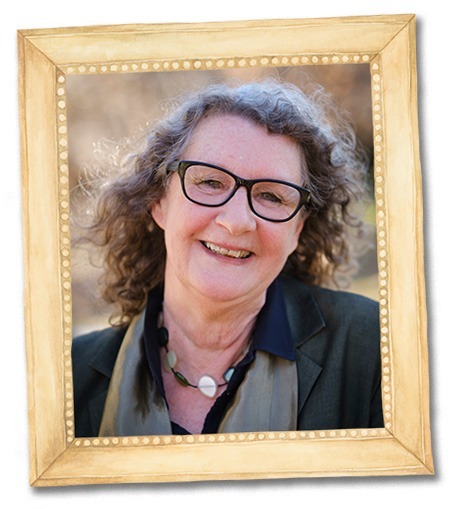 A smiling woman with curly hair and glasses, wearing a dark top, necklace, and a green scarf, is pictured in a wooden frame with a beaded border—a perfect portrait for the Peculiar Parents collection. The background is softly blurred.