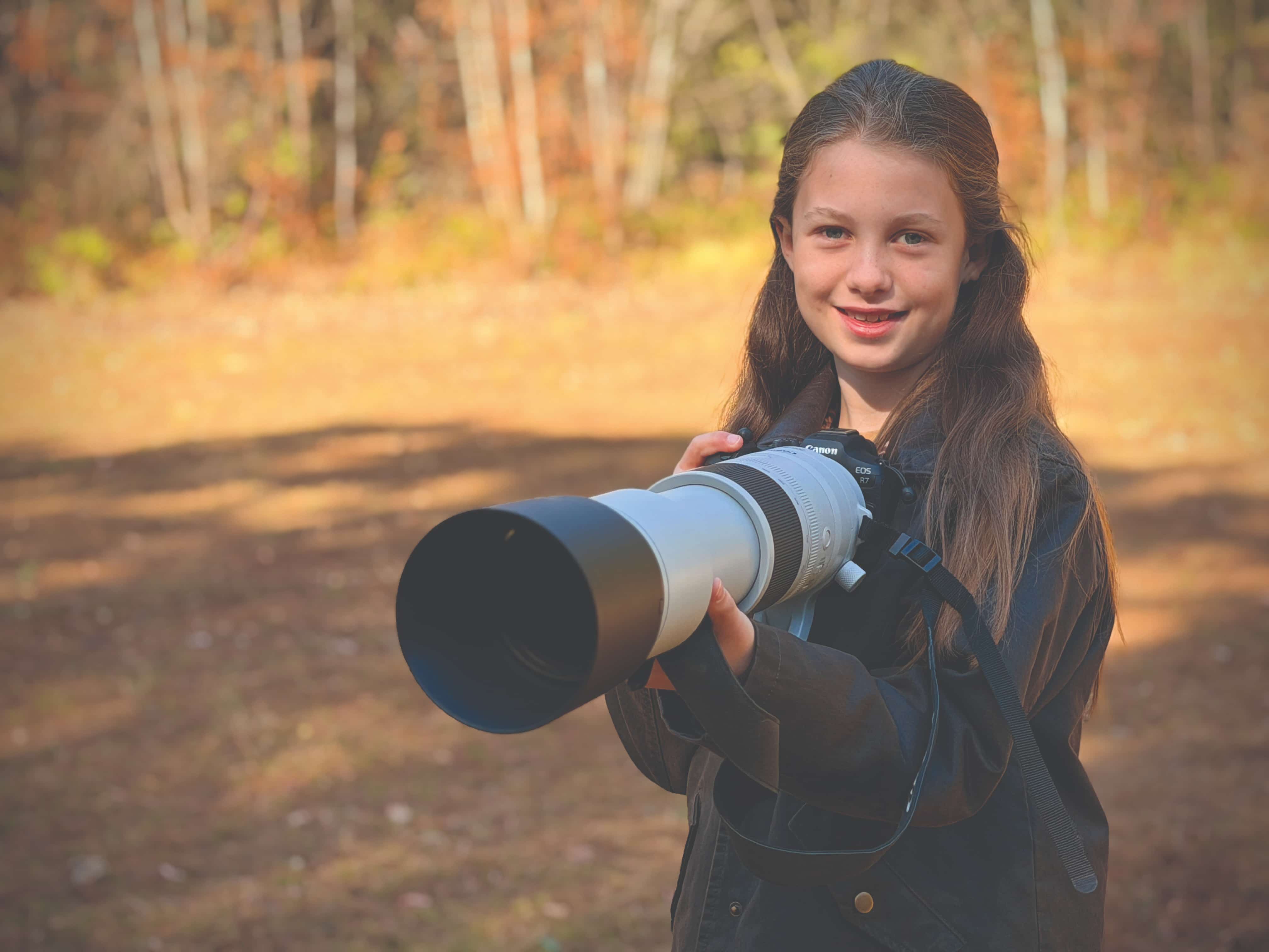 A young girl working as a photographer whilst holding a camera with a long telephoto lens outdoors.