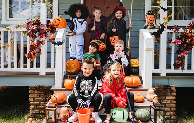 Hallowen facts | a group of kids sit on the steps at the front of a house, all dressed in Halloween costumes. They are holding pumpkins and the house is decorated with autumn leaves and paper bats.