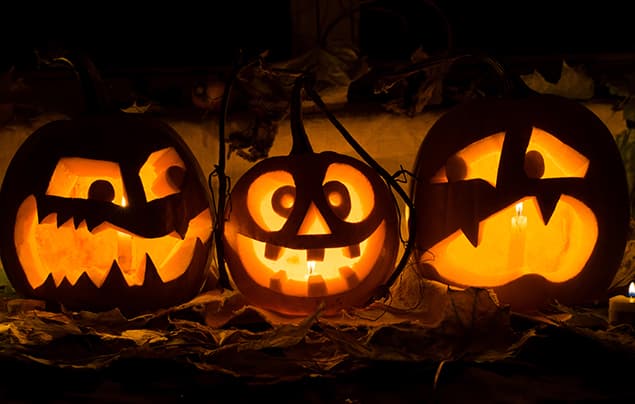three carved vegetables sit in a row. The one on the far left has a scary-looking carved face, the one in the middle has an innocent grin, and the one on the far right looks a little scared! They all have candles burning inside them.