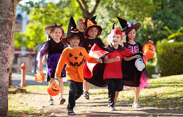 Halloween facts | a group of children head outside to go trick or treating. They are wearing pointy witches' hats and a variety of costume items including a red cloak and an orange t-shirt with a scary face on it like one you'd find on a carved pumpkin.