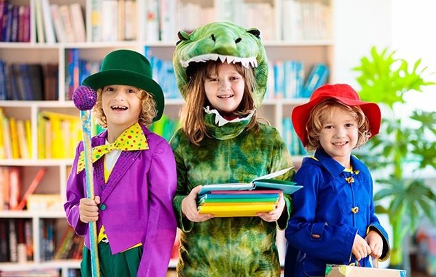 three kids stand in a library dressed in outfits inspired by book characters. A boy is dressed in a purple coat and green hat, a girl wears a crocodile onesie and a boy is dressed as paddington in a red hat and blue jacket