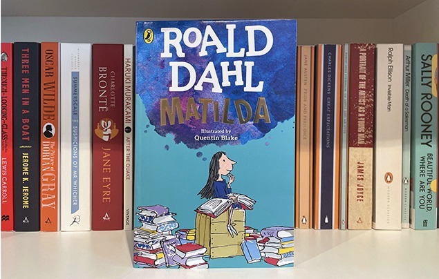 a copy of Matilda by Roald Dahl leans against a shelf of classic books. The cover shows a young girl wearing a blue dress sat on top of a chest of drawers. She is surrounded by books.