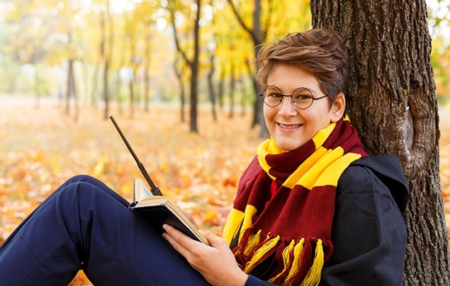 World Book Day | a young boy leans against a tree dressed as Harry Potter. He's wearing a red-and-yellow scarf, school uniform, glasses and is holding a wand and balancing a book on his knees