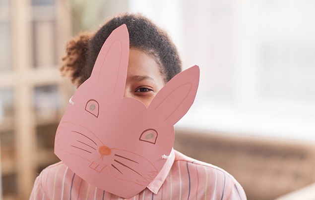 a young girl holds up a homemade mask in front of her face - it's a rabbit drawn on pink paper and held on a lollipop stick. She is wearing pink to match.