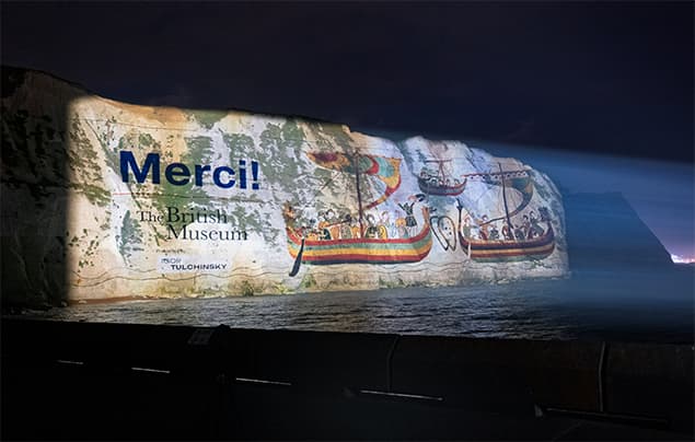 A night projection on white cliffs shows “Merci! The British Museum”, with colourful ancient ships and figures, illuminated above the sea.