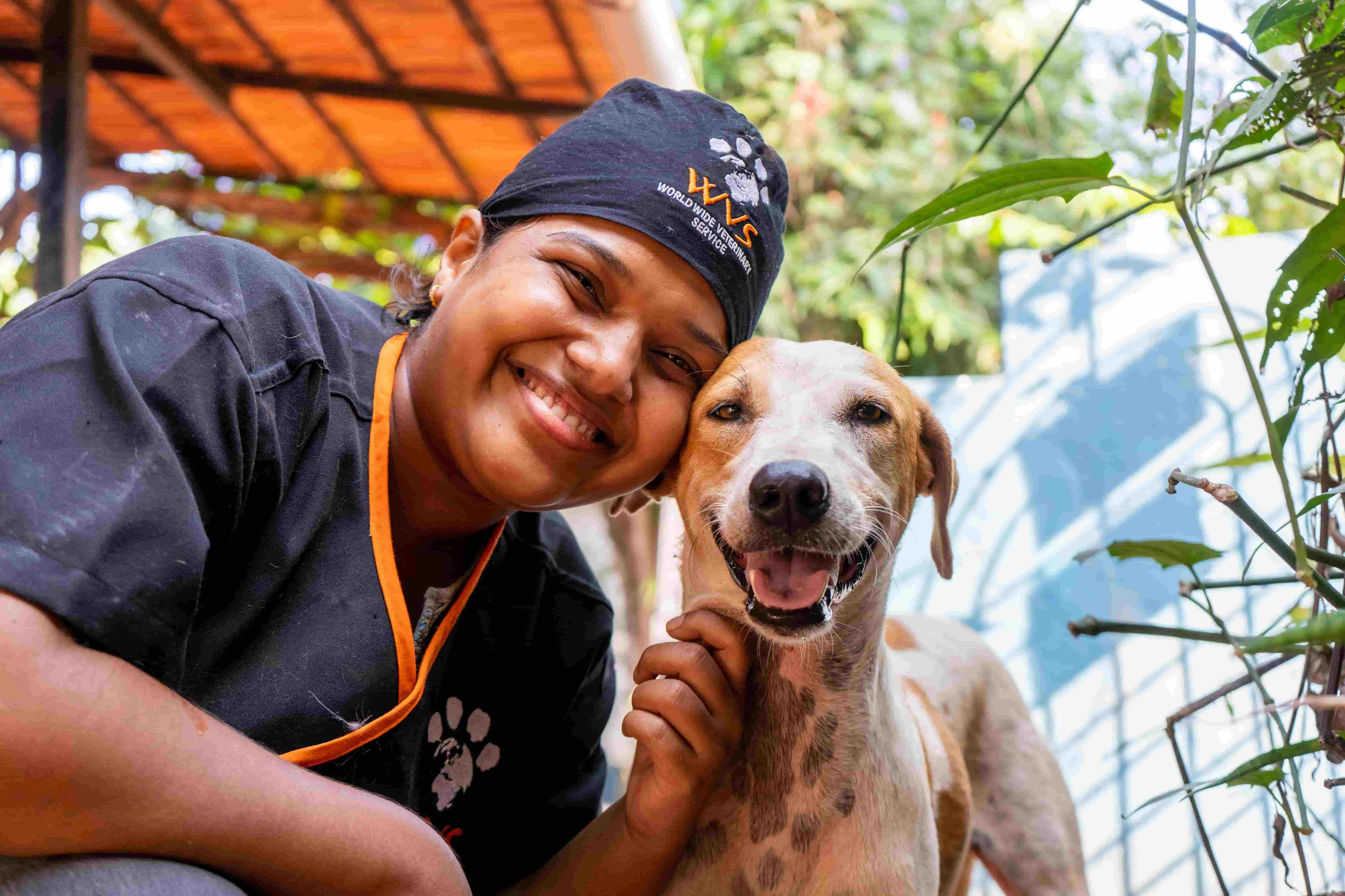 A smiling member of the Young Vets' Club in veterinary scrubs and cap poses outdoors with a happy brown and white dog, both looking at the camera.