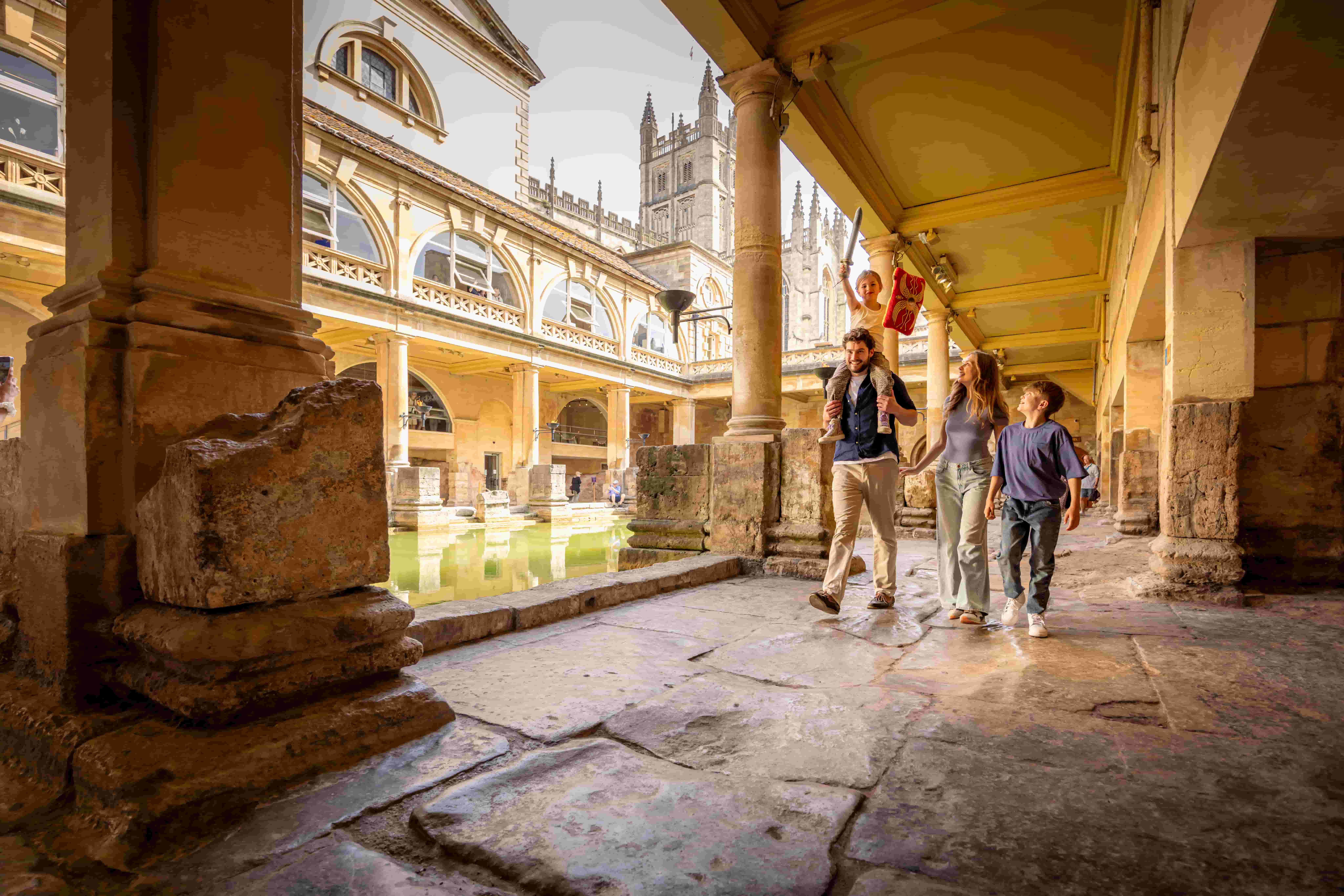 A family of three walks through ancient stone columns beside a reflective pool at the Roman Baths in Bath, England, surrounded by historic architecture and with a church visible in the background.