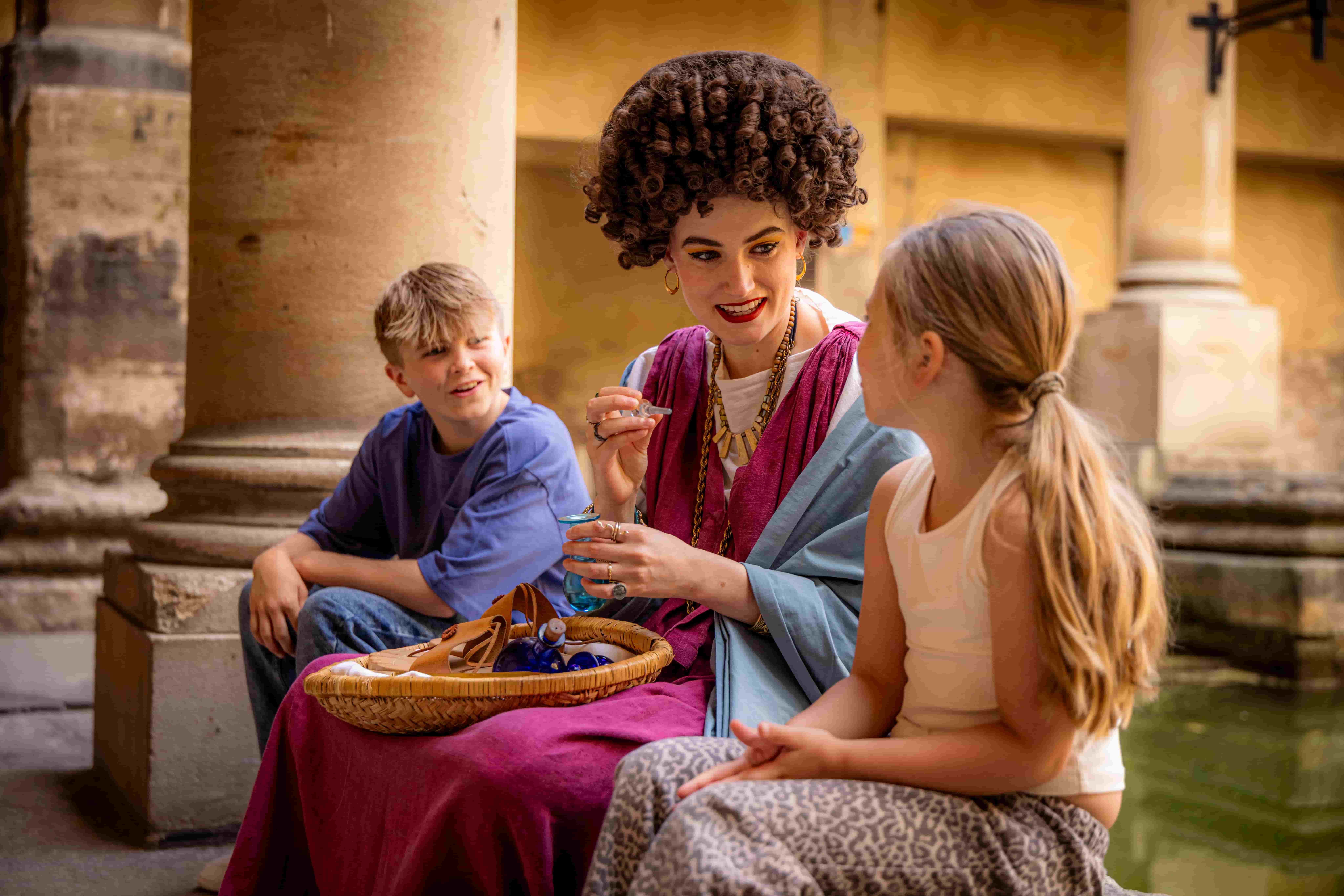 A woman in historical clothing sits between a boy and a girl. She holds a small object and smiles, while the children watch her. They are sitting by a shallow pool surrounded by stone columns, evoking the tranquil atmosphere of Roman Baths.