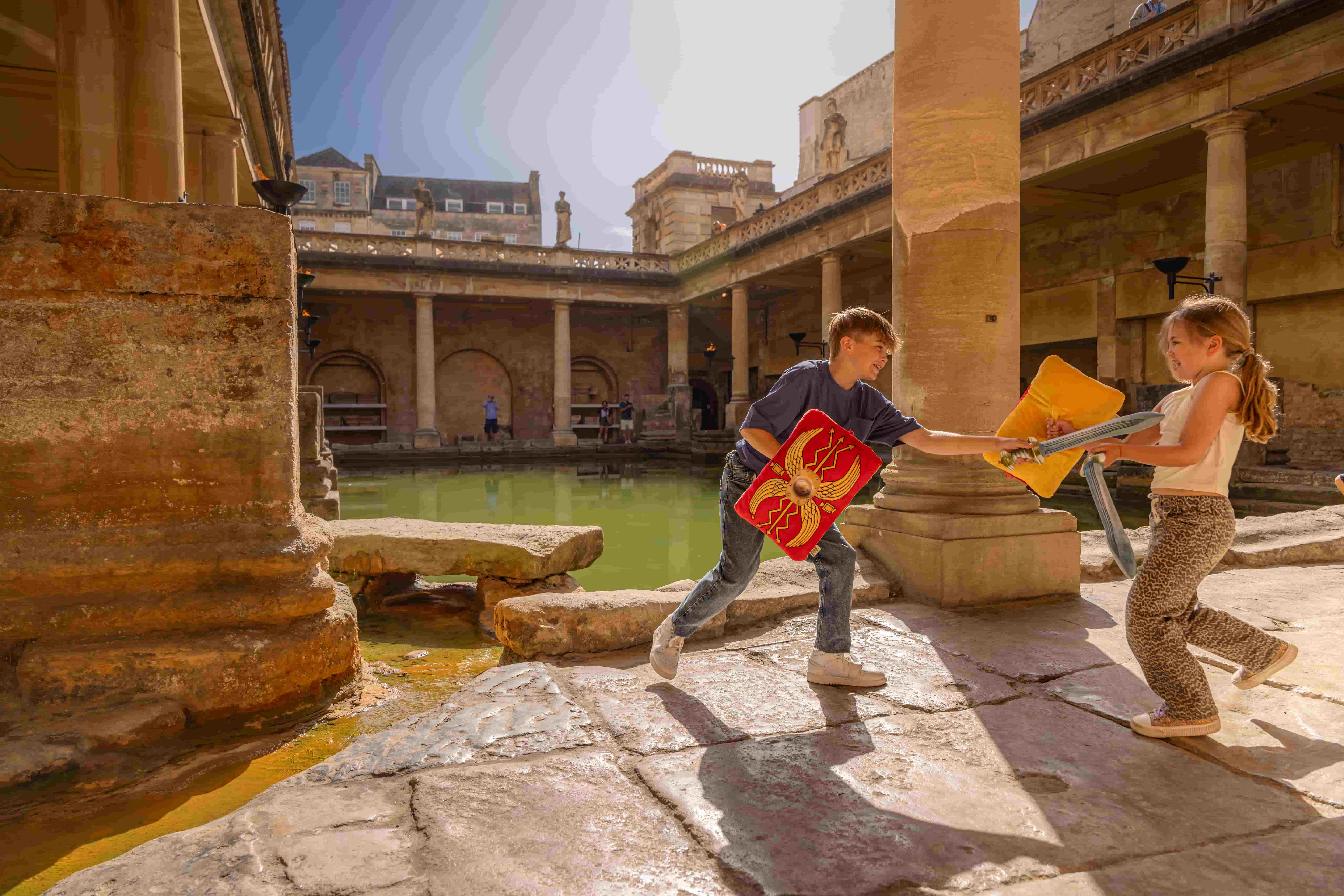 Two children playfully sword fight with foam swords and shields beside the Roman Baths, sunlight streaming onto the stone courtyard and greenish water in the centre—a lively scene echoing the rich history of ancient Roman sites in Bath, England.