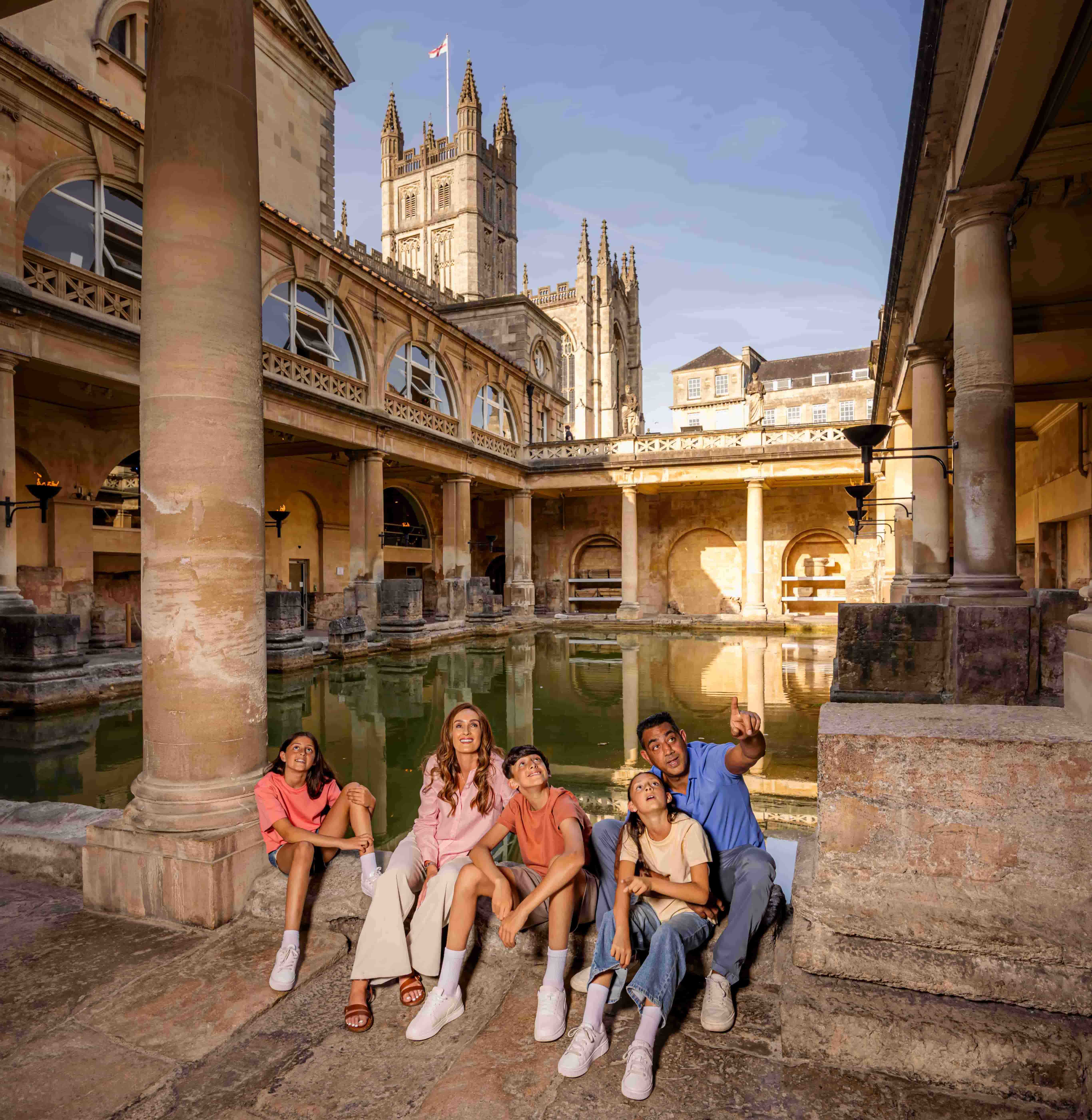 Five people sit and smile by the Roman Baths in Bath, UK, with historic columns, arched windows, and the Bath Abbey tower visible in the sunny background.