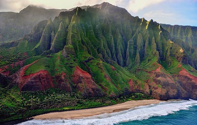 Aerial view of Hawaii’s Na Pali Coast with dramatic green cliffs, red earth, sandy beach, and blue ocean waves under a partly cloudy sky—a breathtaking landscape that’s as surprising and delightful as the best knock knock jokes.