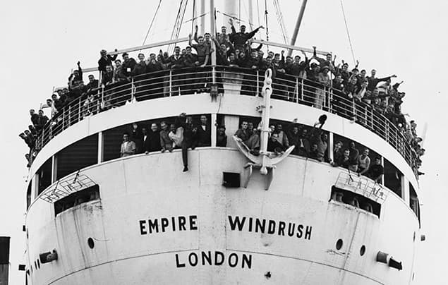 Black and white photo of a crowded ship, the Empire Windrush, with people smiling and waving from the deck. Marking the beginning of the Windrush Generation, the ship’s name, EMPIRE WINDRUSH LONDON, is visible on the bow.