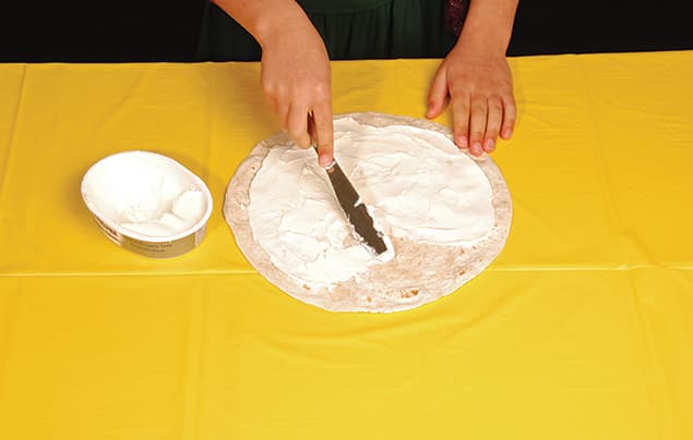 A person spreads a white, creamy substance onto a flat tortilla with a knife on a yellow tablecloth, preparing the base for a colourful rainbow wrap. A tub of the spread sits beside the tortilla.