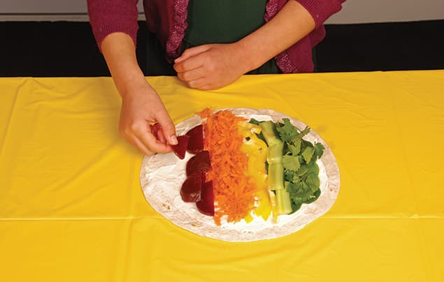 A person arranges sliced vegetables in colourful rows on a tortilla, creating a vibrant rainbow wrap on a yellow tablecloth.