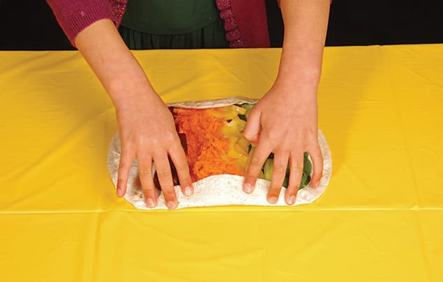 A person is rolling a rainbow wrap filled with shredded carrots, lettuce, and other vegetables on a yellow tablecloth.
