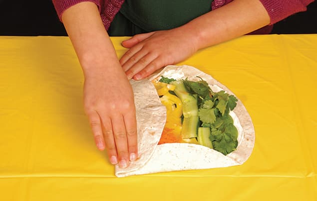A person rolling a vibrant rainbow wrap filled with vegetables and herbs on a yellow tablecloth.