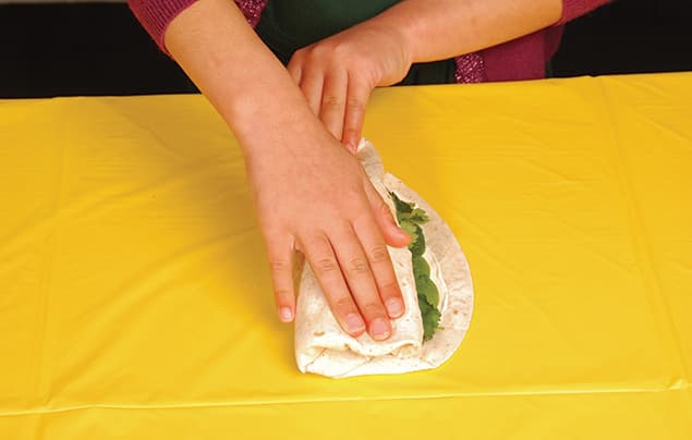 A person folding a rainbow wrap filled with fresh greens on a yellow tablecloth.