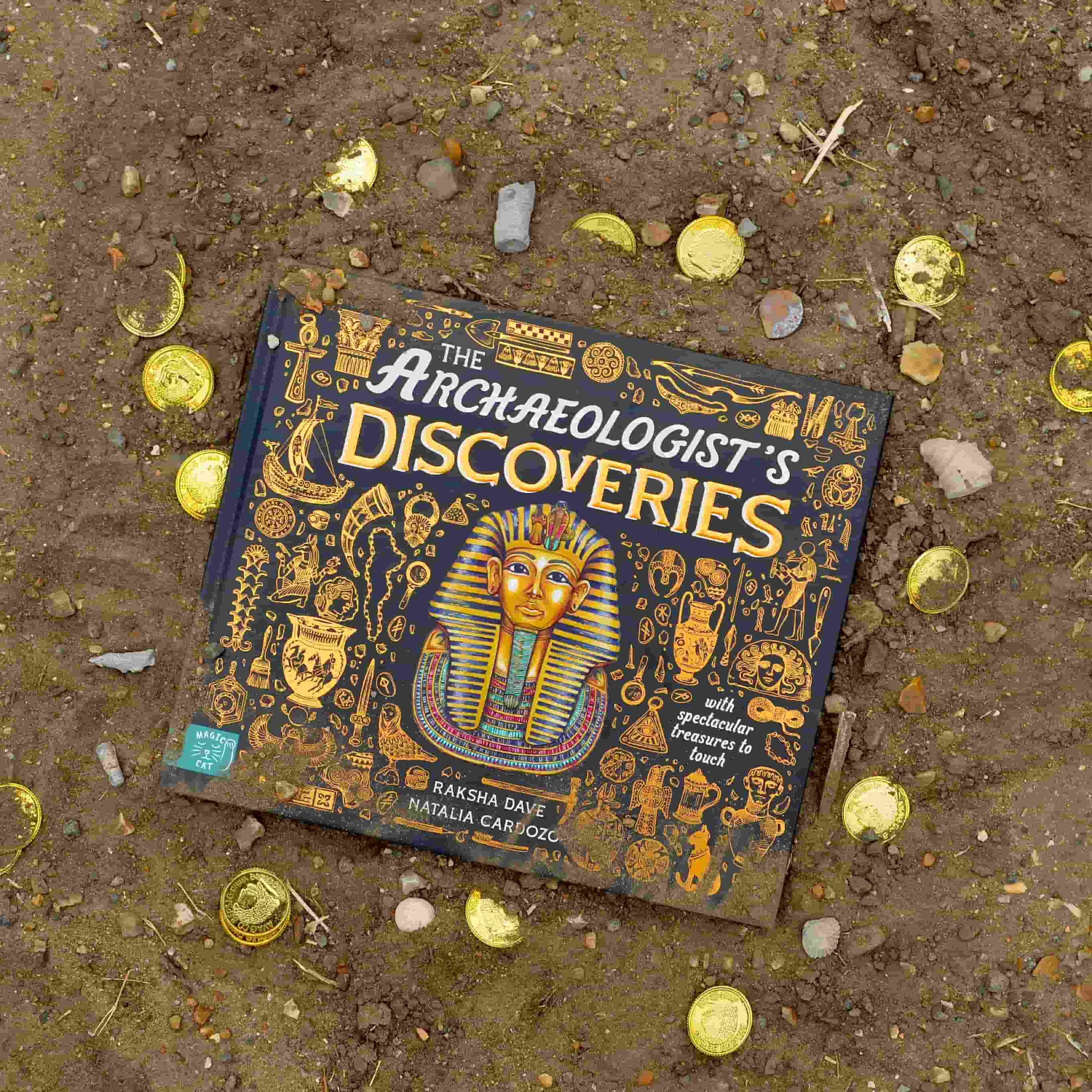 A book titled The Archaeologist’s Discoveries lies on soil, surrounded by scattered gold coins. The cover features Egyptian-themed illustrations and a pharaoh’s mask.