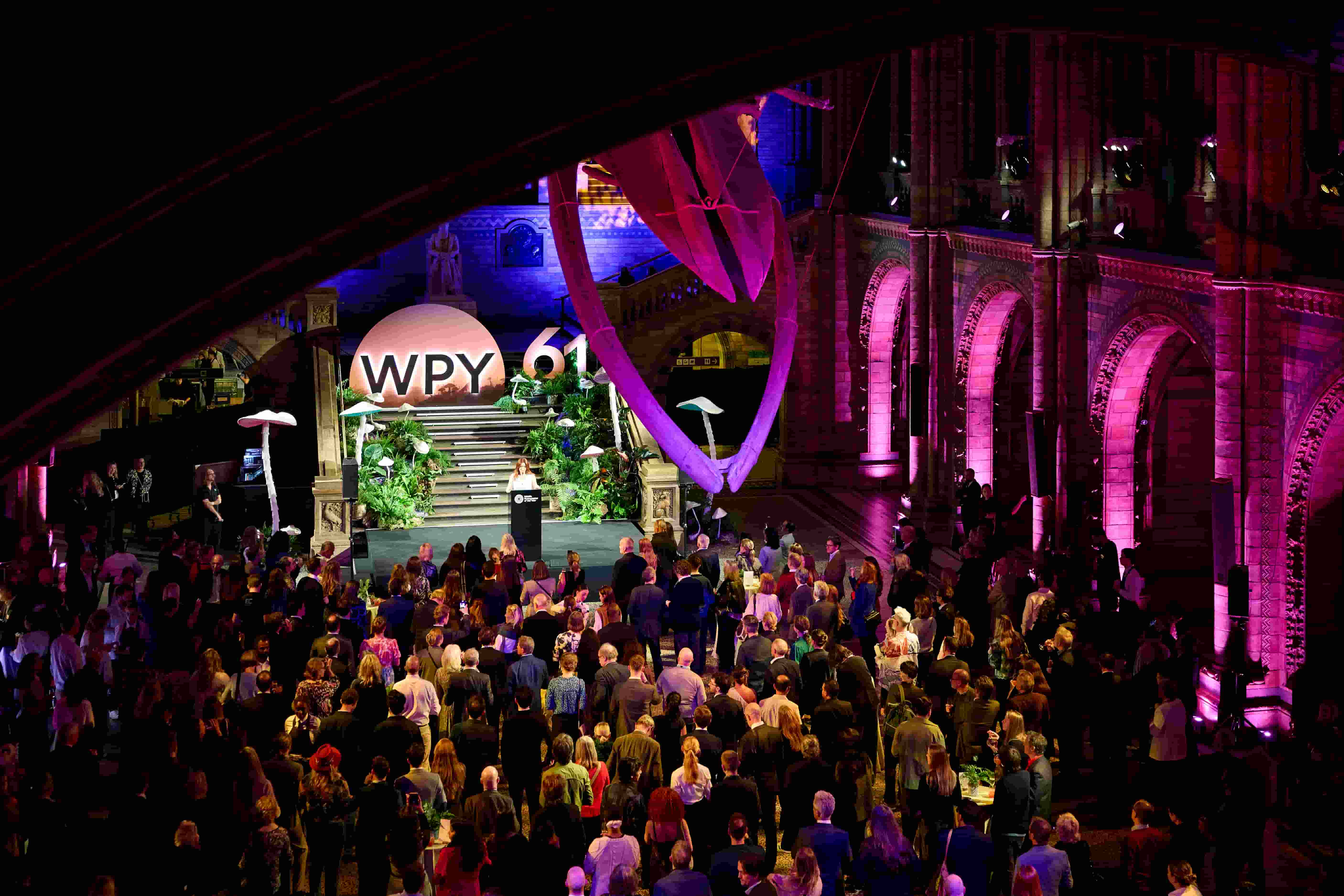 A person stands at a lectern on a stage in a large hall during the Wildlife Photographer of the Year 61 awards ceremony, facing a crowd of attendees at the photography competition.